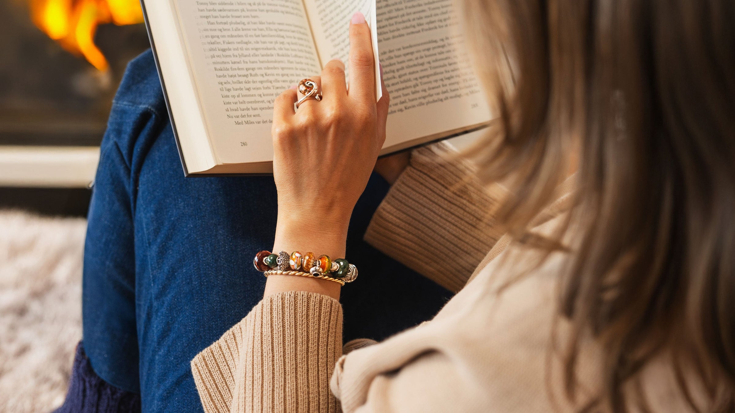Person reading a book with a warm, cozy background while wearing Trollbeads Hygge collection in bracelet and ring with gold plated bangle.